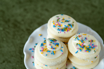 Three stacks of vanilla frosted sugar cookies with multi-color sprinkles on a white cake plate, on a green background