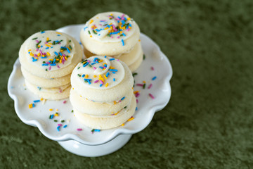 Three stacks of vanilla frosted sugar cookies with multi-color sprinkles on a white cake plate, on a green background