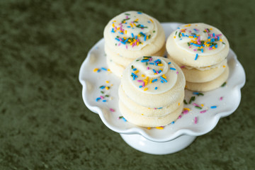 Three stacks of vanilla frosted sugar cookies with multi-color sprinkles on a white cake plate, on a green background