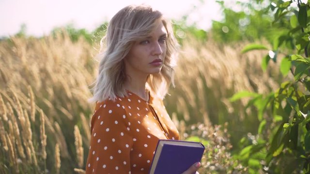 Beautiful happy young woman in field of spikelets and wheat with book on the sunset, blonde in the grass having a good time