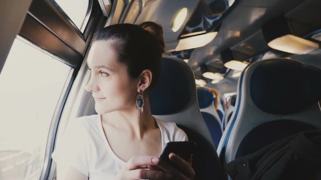 Close-up view of beautiful happy smiling Caucasian girl using smartphone, looking out of train window and walking away.