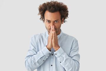 People and faith concept. Handsome young serious African American male keeps hands in praying gesture, looks confidently at camera, dressed in white shirt, has strong believe his dreams come true.