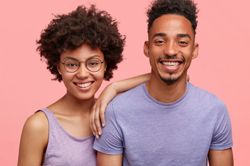 Horizontal shot of happy African American woman and man have truthful relationships, toothy smile, happy to meet with friends, dressed casually, isolated over pink background. Emotions concept