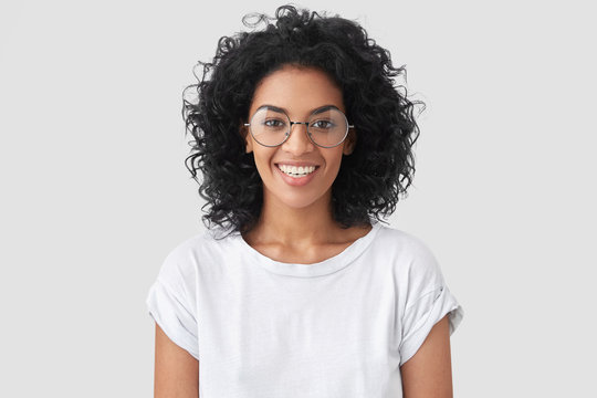 Beautiful smiling African American female with crisp hair, broad smile, shows white teeth, wears casual t shirt and spectacles, stands over studio wall rejoices having day off. Woman journalist indoor