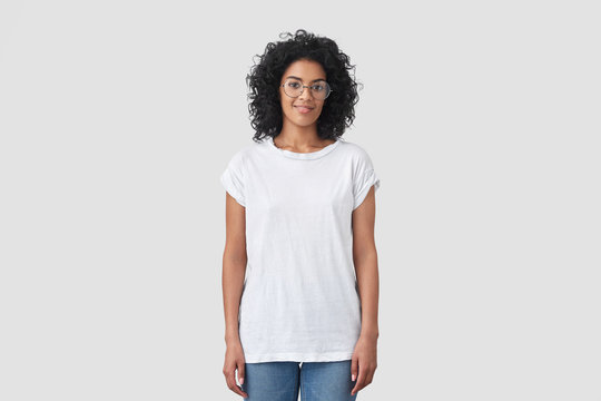 Indoor shot of attractive dark skinned female with crisp hair, wears round glasses, dressed in casual white t shirt and jeans, poses indoor, has Afro hairstyle. Woman student has rest after classes