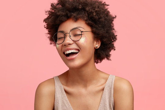 Close up portrait of happy African American female laughs at something funny, has positive expression, wears glasses, has curly hair, dressed casually, closes eyes with happiness, isolated on pink