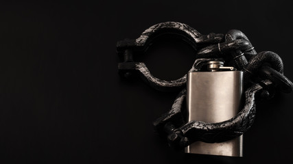 Addiction, alcoholism and alcohol abuse concept with close up on a metal flask containing an alcoholic drink shackled in iron cuffs on a black background with dark dramatic lighting with copy space