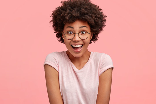 People and reaction concept. Happy overjoyed young African American female reacts on positive news, has broad smile and surprised expression, wears casual t shirt, poses against pink background