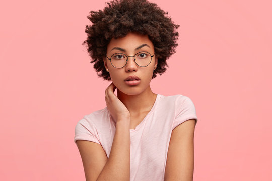 Adorable serious African American female with self assured expression, keeps hands on neck, dressed in casual shirt in one tone with background, has healthy skin, models indoor. People and ethnicity
