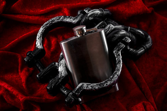 Addiction, alcoholism and alcohol abuse concept with close up on a metal flask containing an alcoholic drink shackled in iron cuffs on a red velvet background with dramatic dark lighting