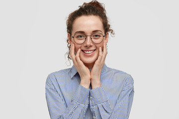 Positive satisfied young woman has freindly gentle smile, touches cheeks with hands, dressed in stylish shirt, happy to have date with boyfriend and recieve present, poses over white studio wall