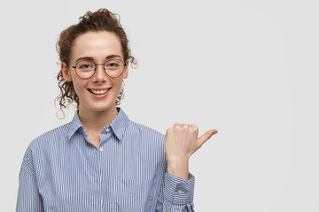 Lovely freckled attractive young woman with joyful expression, has freckles on skin, dressed in stylish shirt, points aside with thumb, shows free space for advertisement or promotional content.