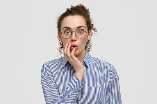 Image of impressed stunned freckled female hears surprising news, keeps hand on cheek, dressed in striped shirt, stands over white background. Astonished amazed young woman with bated breath