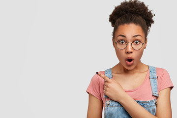 Image of emotive young African American woman with stupefied expression, dropps jaw while points left, demonstrates stunning sale, poses against white background, wears round spectacles and dungarees