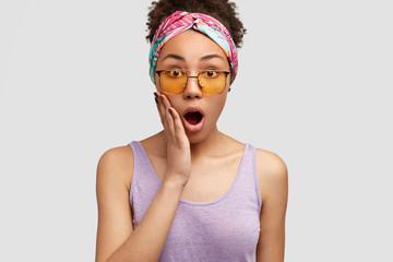 Photo of emotive surprised Afrcan American female opens mouth widely, keeps palms on cheek, wears stylish headband and purple vest, isolated over white background. People and emotions concept