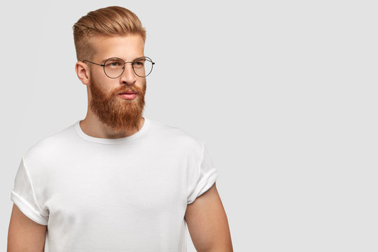Brutal young handsome man with ginger beard, trendy hairstyle, wears casual white t shirt and spectacles, looks aside, stands against white background with free space for your advertisement.