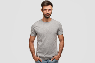 Horizontal portrait of attractive bearded male with serious expression, dressed in casual grey t shirt, keeps hands in pockets, shows new clothes, isolated over white background. People, style concept
