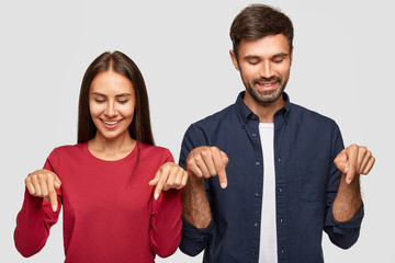 Positive young girlfriend and boyfriend with joyful expression, notice something pleasant down,...