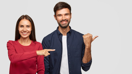 Cheerful brunette young female in red sweater with healthy skin, positive bearded guy stand together in studio, point aside at blank space, show place for your advertisement or promotional text