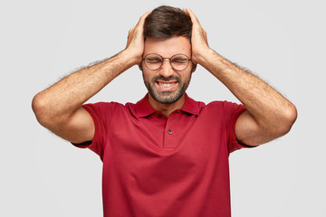 Fototapeta premium Horizontal shot of fatigue young Euroepean unshaven male keeps hands on head, feels exhausted after work in office, has headache, wears casual bright red t shirt, isolated over white background