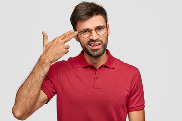 Fototapeta premium Young stressful male makes gun gesture, pretends commiting suicide, keeps two fingers on temples, feels desperate, poses alone against white background, dressed in casual red t shirt, has crisis