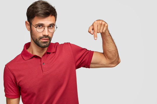 Confident unshaven male model in eyewear and red t shirt, points down, attracts your attention to blank space for advertisement or promotional text, isolated on white studio wall. Lifestyle concept