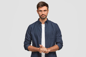 Waist up portrait of handsome serious unshaven male keeps hands together, dressed in dark blue shirt, has talk with interlocutor, stands against white background. Self confident man freelancer