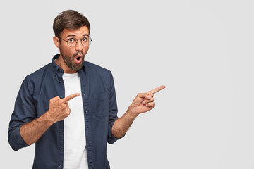 Impressed Caucasian male with stubble, retells gossip to friend, points right, being impressed and shocked, dressed in fashionable shirt, stands against white blank wall for your promotional content