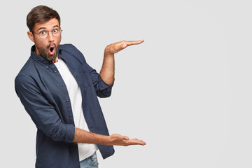 Stunned unshaven male with shocked facial expression gestures with hands, shows size or height of...