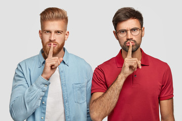 Two male friends with thick beards, keep fore fingers on lips, look secretly at camera, tell very...