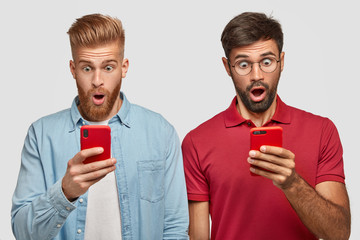Surprised two male student being addicted to modern electornic devices, hold smart phones, stare at...