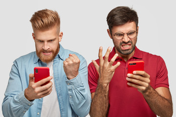 Irritated two guys look angrily at screen of smart phones, watch football match online, being...