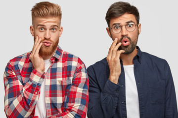 Bewildered two male companions look with puzzled expressions, gaze at something awesome, keep hands...