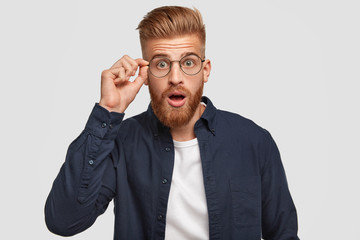Stupefied hipster guy with shocked expression, hears something unbelievable, keeps hand on rim of...