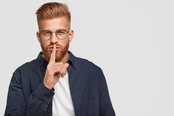Secret ginger young man has mysterious expression, touches lips with fore finger, dressed casually, poses against white background with copy space for your advertisement or promotion. Hush sign