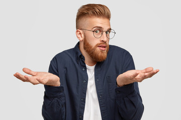 Photo of confused bearded ginger male in spectacles looks surprisingly at camera, shruggs shoulders...