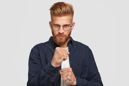 People and power concept. Serious foxy man with determined expression, keeps clenched fists, looks with stern at camera, ready to fight, dressed in dark blue shirt, stands against white wall