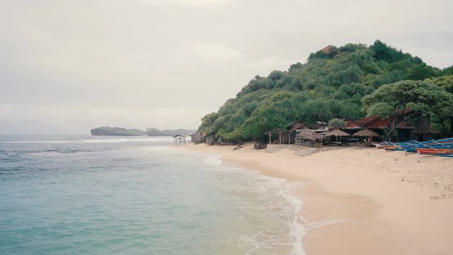 White sandy beach with straw umbrellas and houses near green hill in Java, Indonesia
