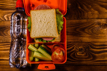 Bottle of water and lunch box with sandwich, cucumbers and tomatoes on wooden table. Top view