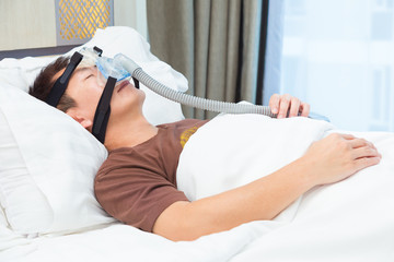 Obraz na płótnie Canvas Asian man wearing CPAP mask, connecting to air hose, sleeping on his bed in his bedroom