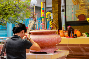 An Asian and elderly woman is praying in a Chinese temple at the altar to the gods and spirits with incense sticks for luck, health and prosperity. She has a strong faith and belief.