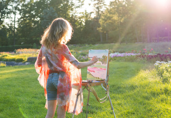 A young woman artist holds a brush and paints a picture on an easel in the rays of the sunset.