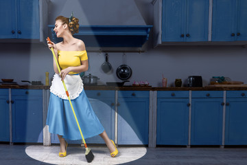 Retro pin up girl woman female housewife wearing yellow top, blue skirt and white apron holding mop singing and cleaning floor in stage light kitchen with blue cabinets and utensils. Housework concept