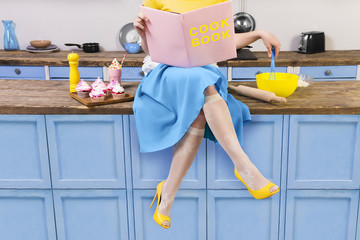 Colorful retro / pin up girl woman female housewife wearing yellow top, skirt and white apron sitting on kitchen and reading pink cook book. Sweet food cupcakes and milkshake home cooked on the table