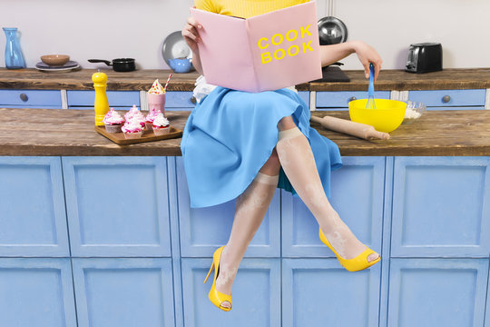 Colorful retro / pin up girl woman female housewife wearing yellow top, skirt and white apron sitting on kitchen and reading pink cook book. Sweet food cupcakes and milkshake home cooked on the table