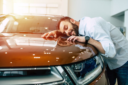 Happy handsome bearded man buying a car in dealership, guy hugging hood of new car