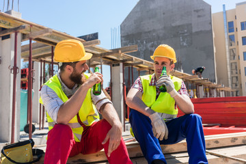 Two young workers smiling while drinking a cold alcoholic or non-alcoholic beer, during break at...