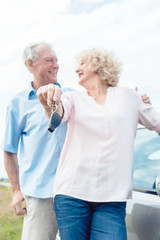 Close-up of the hands of a senior woman showing the keys of her car while posing next to her partner as confident elderly drivers