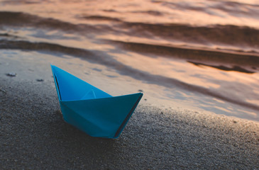 Paper blue ship is thrown on sandy shore of lake at sunset.
