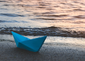 Paper blue ship is thrown on sandy shore of lake at sunset.
